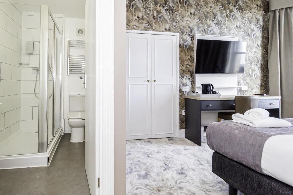 A hotel room with a shower, toilet, wardrobe, desk, and a bed with a TV above it.