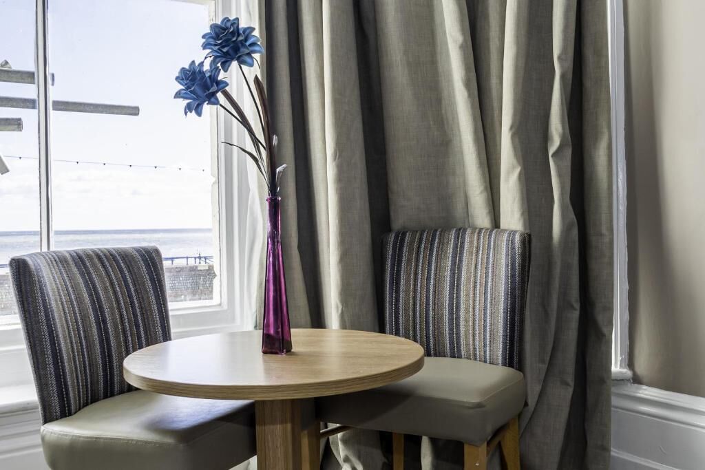 A round wooden table with two upholstered chairs, a purple vase with blue flowers, and a view of the sea through a window with curtains.
