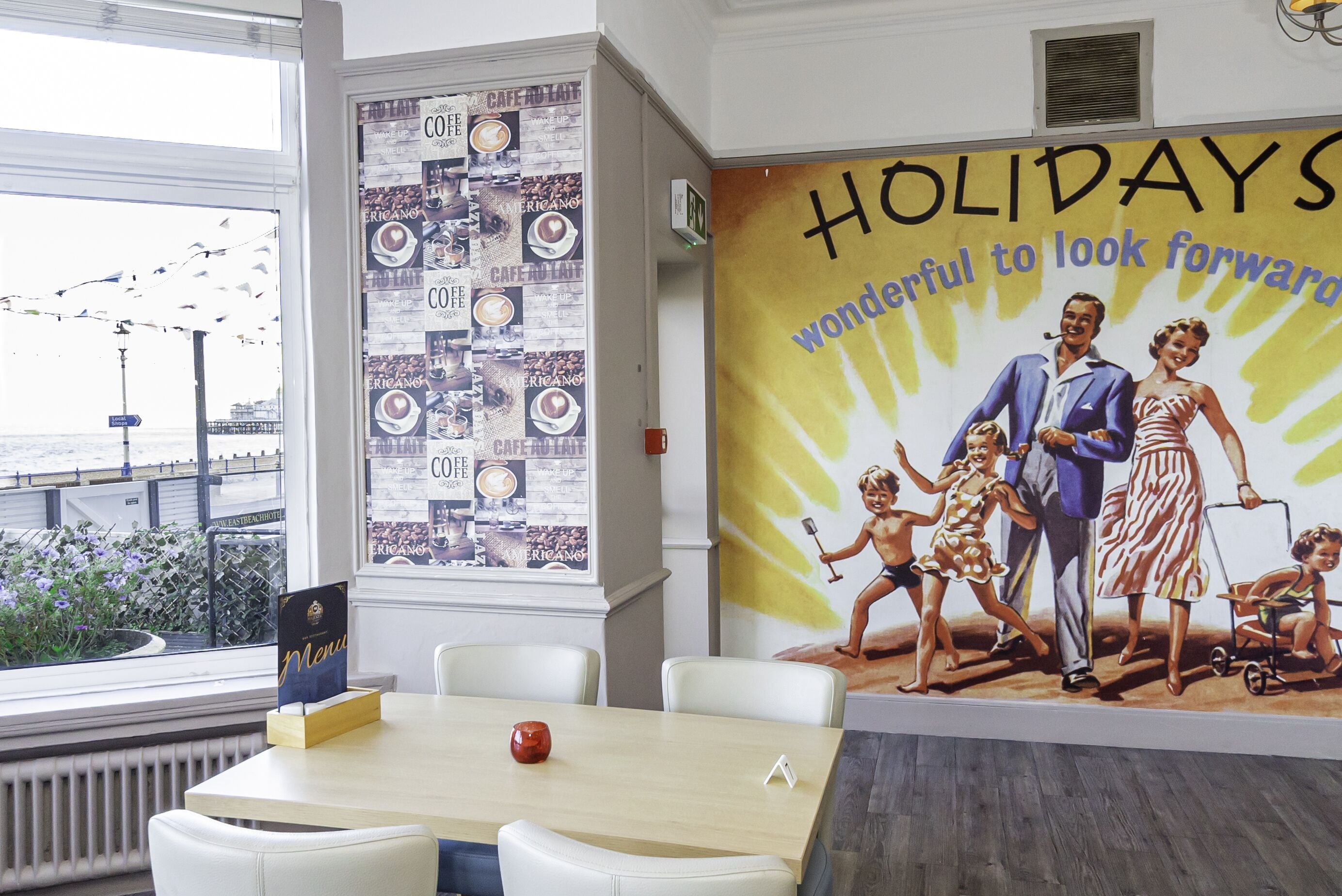 Un espace repas comprenant une table, des chaises, un présentoir à menus, une grande affiche représentant une famille et le texte « LES VACANCES, une période que l’on attend avec impatience », ainsi qu’une vue sur la mer par la fenêtre.