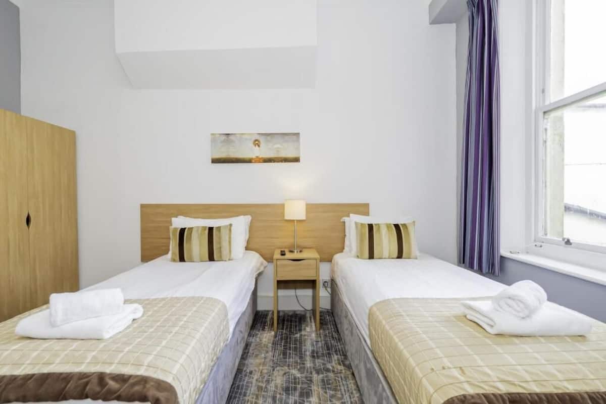 Een hotelkamer met twee bedden, een nachtkastje, een schilderij aan de muur en een raam met gordijnen.