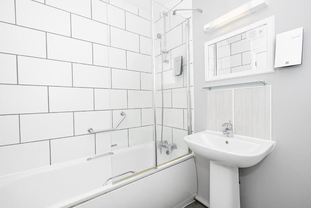 Un baño moderno con azulejos blancos, cabina de ducha de vidrio, lavamanos blanco y un espejo.