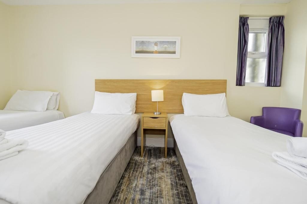 Habitación de hotel con dos camas, cabecera de madera, mesita de noche con lámpara, una silla morada y una ventana con cortinas.