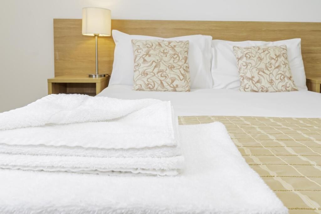 A neatly made bed with white bedding and patterned pillows, a bedside lamp, and a stack of white towels.