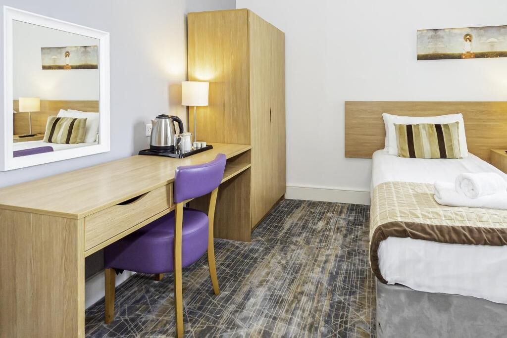 Quarto de hotel com escrivaninha de madeira, cadeira roxa, cama com roupa de cama branca e castanha, e um espelho grande que reflete o quarto.