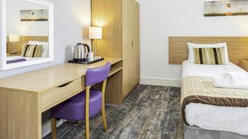 Quarto de hotel com escrivaninha de madeira, cadeira roxa, cama com roupa de cama branca e castanha, e um espelho grande que reflete o quarto.