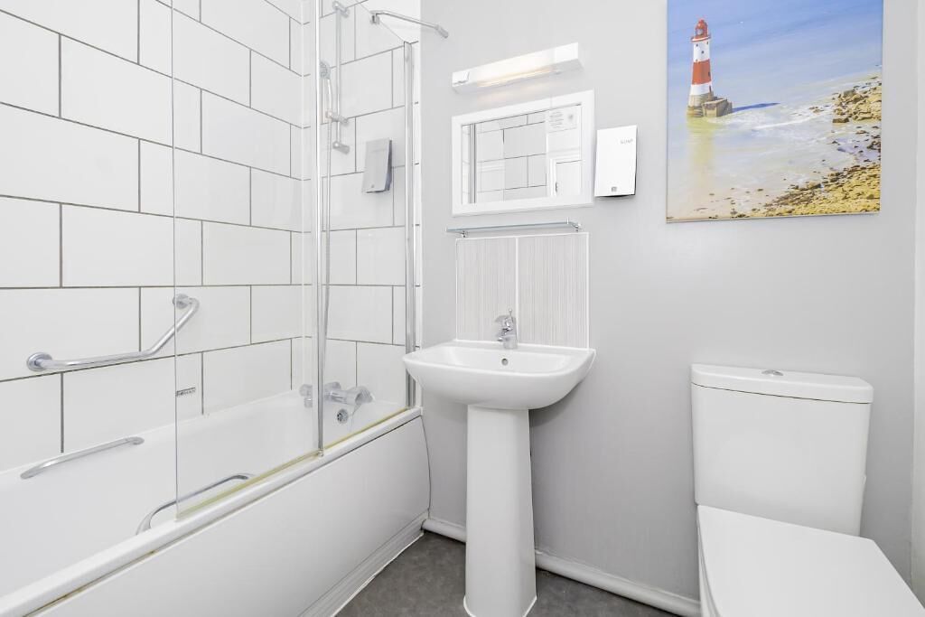 Un baño con baldosas blancas, una bañera con barras de apoyo, un lavamanos pedestal, un inodoro y un cuadro de un faro en la pared.