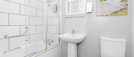 Un baño con baldosas blancas, una bañera con barras de apoyo, un lavamanos pedestal, un inodoro y un cuadro de un faro en la pared.