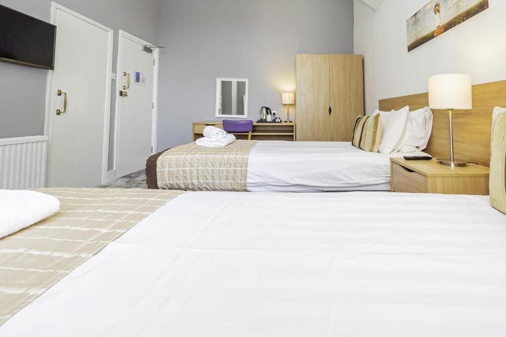 Habitación de hotel con dos camas, un armario de madera, un escritorio con silla, una lámpara y un cuadro en la pared.
