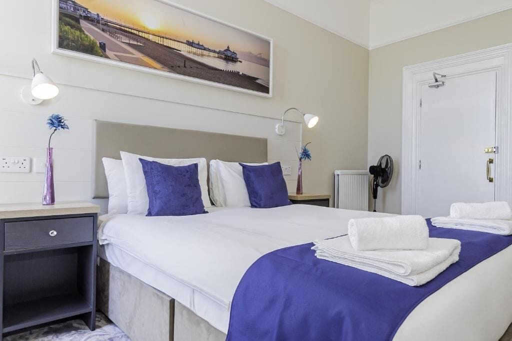 Habitación de hotel ordenada con cama, mesitas de noche, lámpara de pared, radiador y puerta.