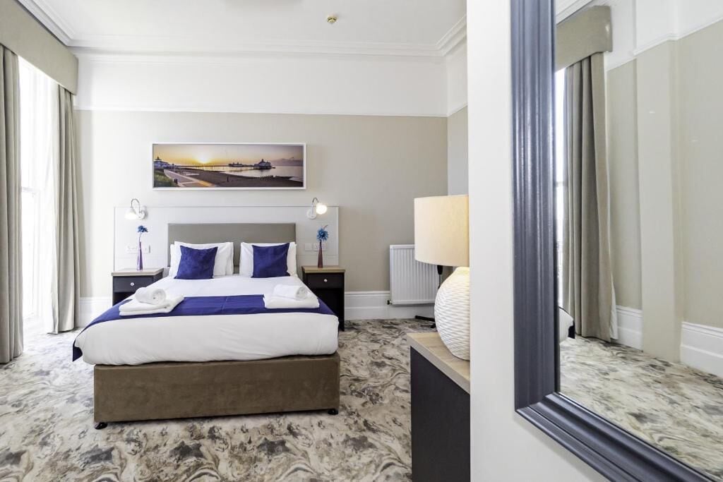 A hotel room with a large bed, a nightstand, a lamp, a painting on the wall, and a mirror.