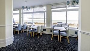 Un espace repas avec des tables et des chaises, de grandes fenêtres équipées de stores et un lustre.