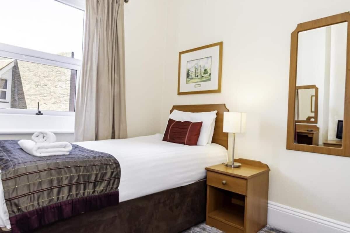 Een hotelkamer met een bed, een nachtkastje met een lamp, een ingelijste foto aan de muur en een raam met gordijnen.