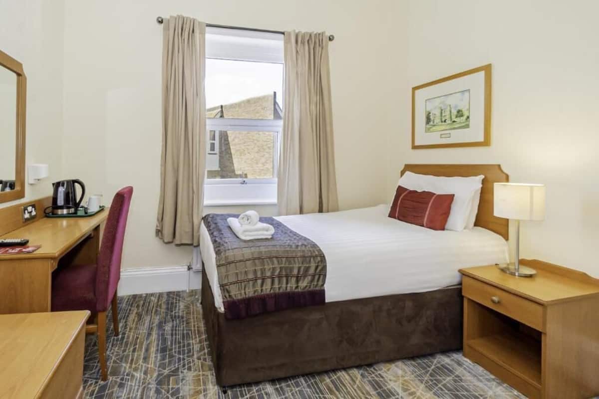 Een hotelkamer met een bed, een bureau, een stoel, een nachtkastje, een lamp, een raam met gordijnen en een ingelijst schilderij aan de muur.