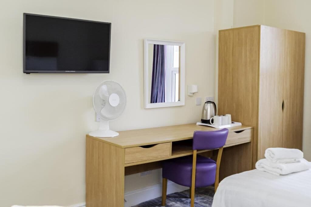 Ein Hotelzimmer mit Holzschreibtisch, violettem Sessel, Wandfernseher, Spiegel und Kleiderschrank.