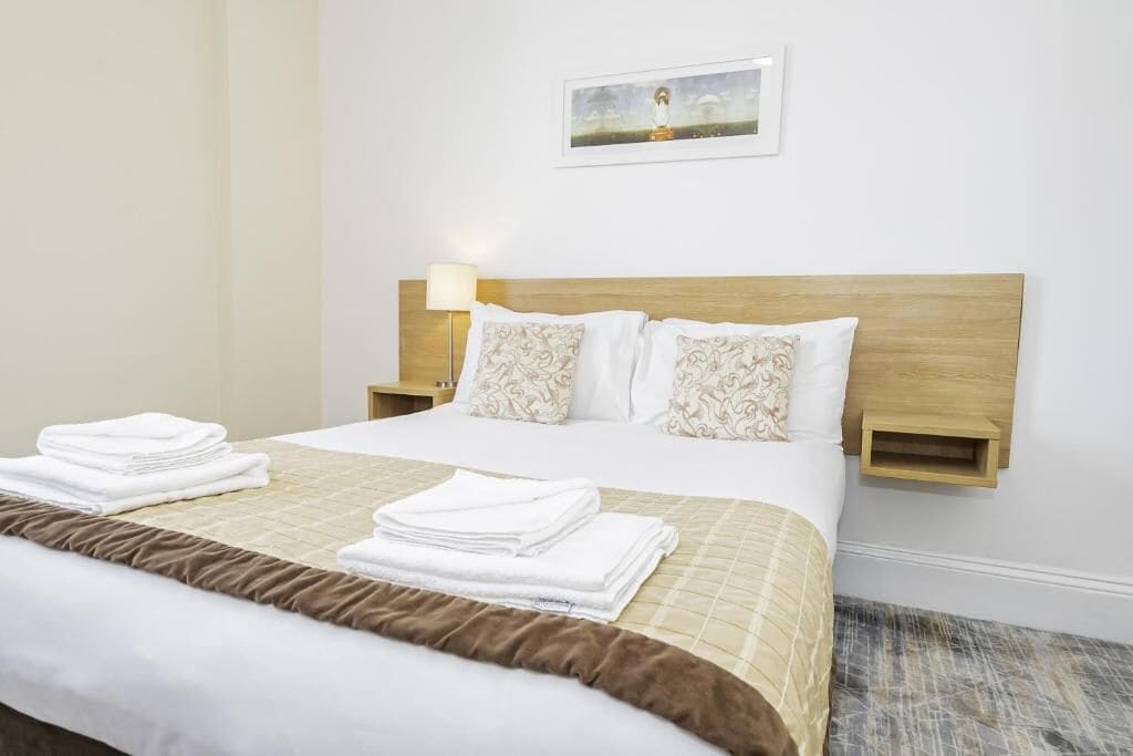 A neatly made bed with a brown accent, white linens, and a wooden headboard. There are two bedside tables with lamps, and a framed picture on the wall.