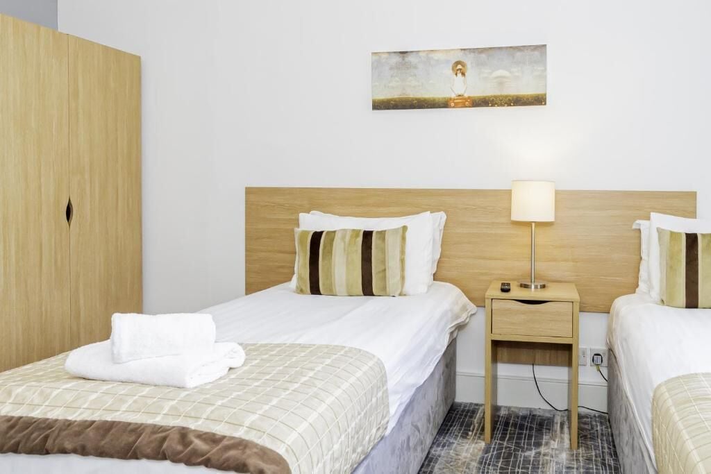 Una habitación de hotel con dos camas, un cabecero de madera, una mesita de noche con una lámpara y un cuadro en la pared.