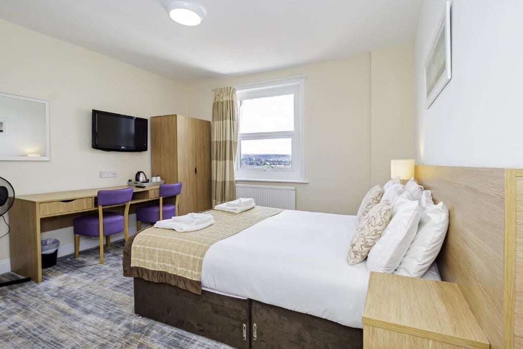 Ein Hotelzimmer mit einem Bett, einem Schreibtisch mit Stuhl, einem Fernseher und einem Vorhangfenster.