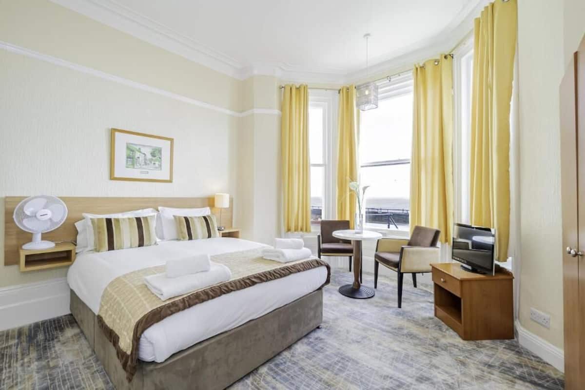 Hotelkamer met een groot bed, een bureau met een televisie, een stoel, een klein tafeltje en een ventilator.