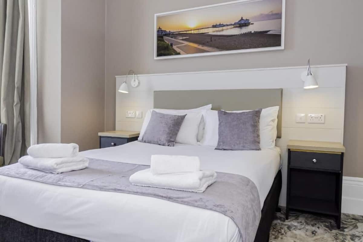 Een net opgemaakt bed met witte en grijze linnens, twee nachtkastjes met lampen, en een schilderij van een zonsondergang boven een pier aan de muur.