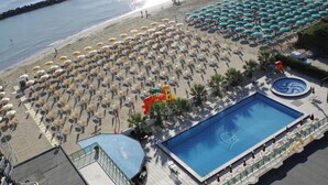 Vista aérea de una playa con numerosas sombrillas y tumbonas, una piscina grande y un litoral rocoso.