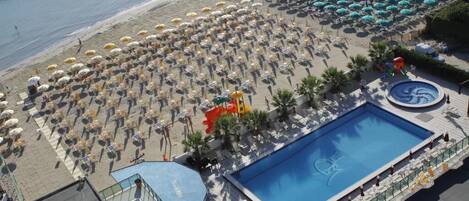 Vue aérienne d’une plage parsemée de nombreux parasols et de chaises longues, avec une grande piscine et une côte rocheuse.