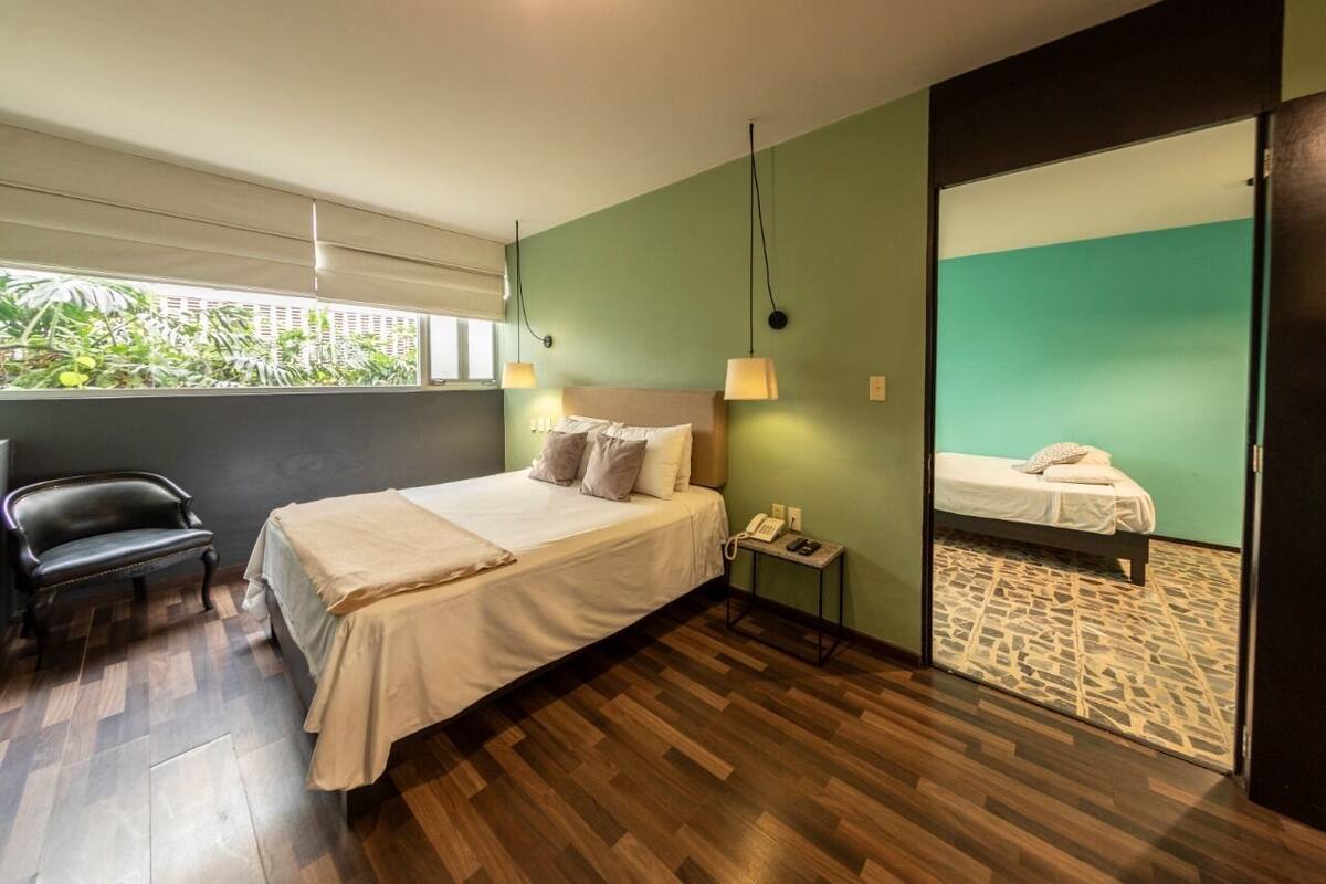 Een moderne hotelkamer met een groot bed, een stoel, een nachtkastje met een telefoon, en uitzicht op een andere kamer door een glazen deur.