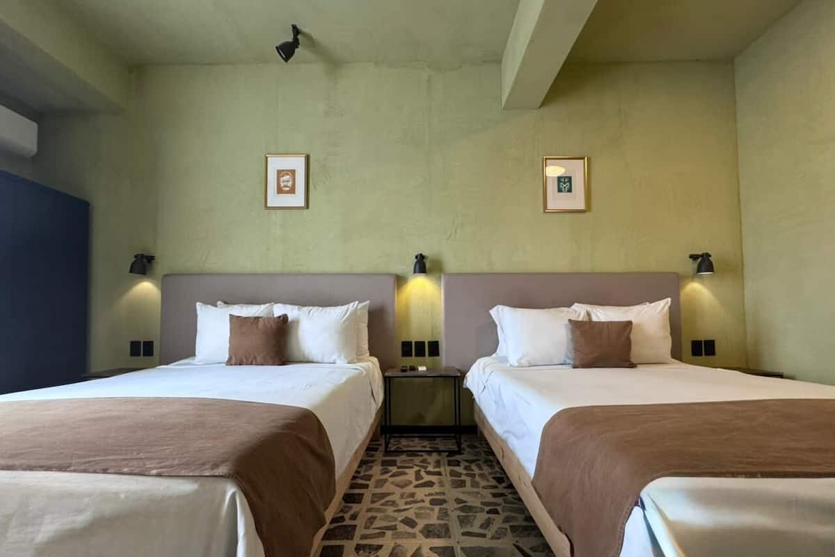Hotelkamer met twee bedden, elk voorzien van witte lakens en bruine plaids. Aan weerszijden van de bedden hangen twee wandlampen, en de vloer is belegd met een betegeling met een patroon.