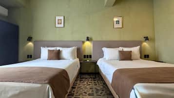 A hotel room with two beds, each with white linens and brown throw blankets. There are two wall-mounted lamps on either side of the beds, and the floor is covered with a patterned tile.