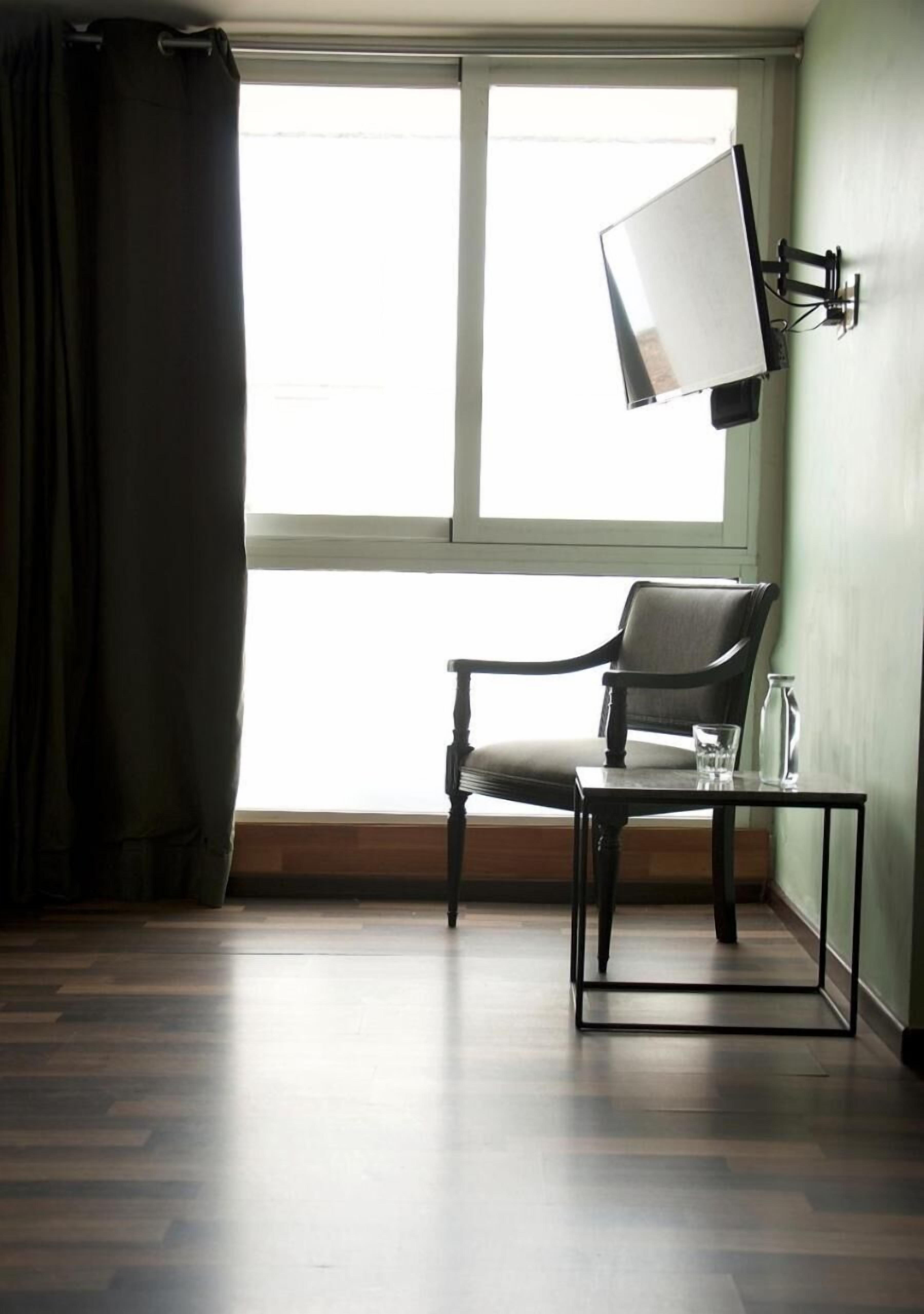 Une pièce avec un plancher en bois, une chaise, une petite table, un téléviseur fixé au mur et une fenêtre avec des rideaux.