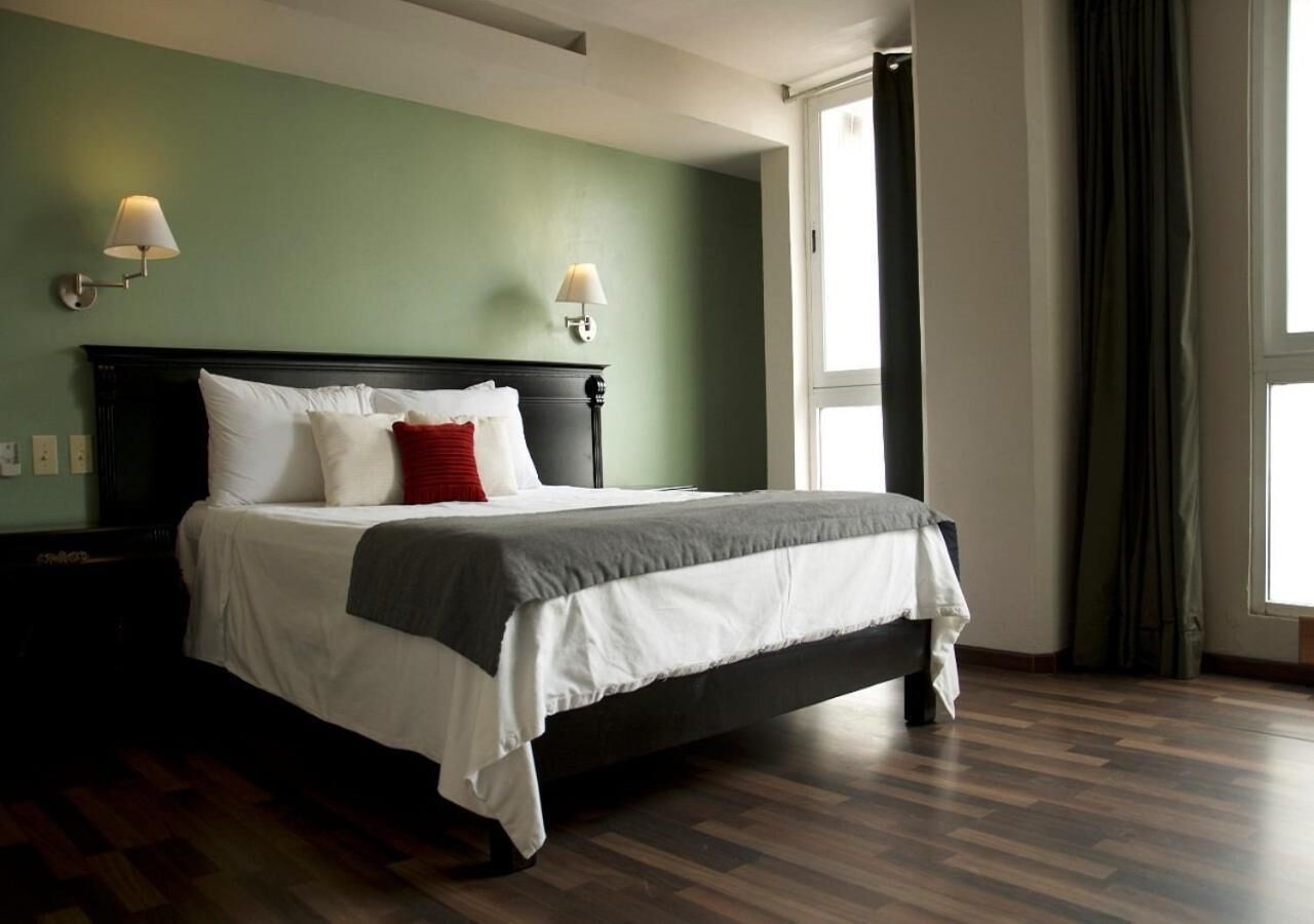 A neatly made bed with white and gray bedding, two wall-mounted lamps, and a large window with curtains.