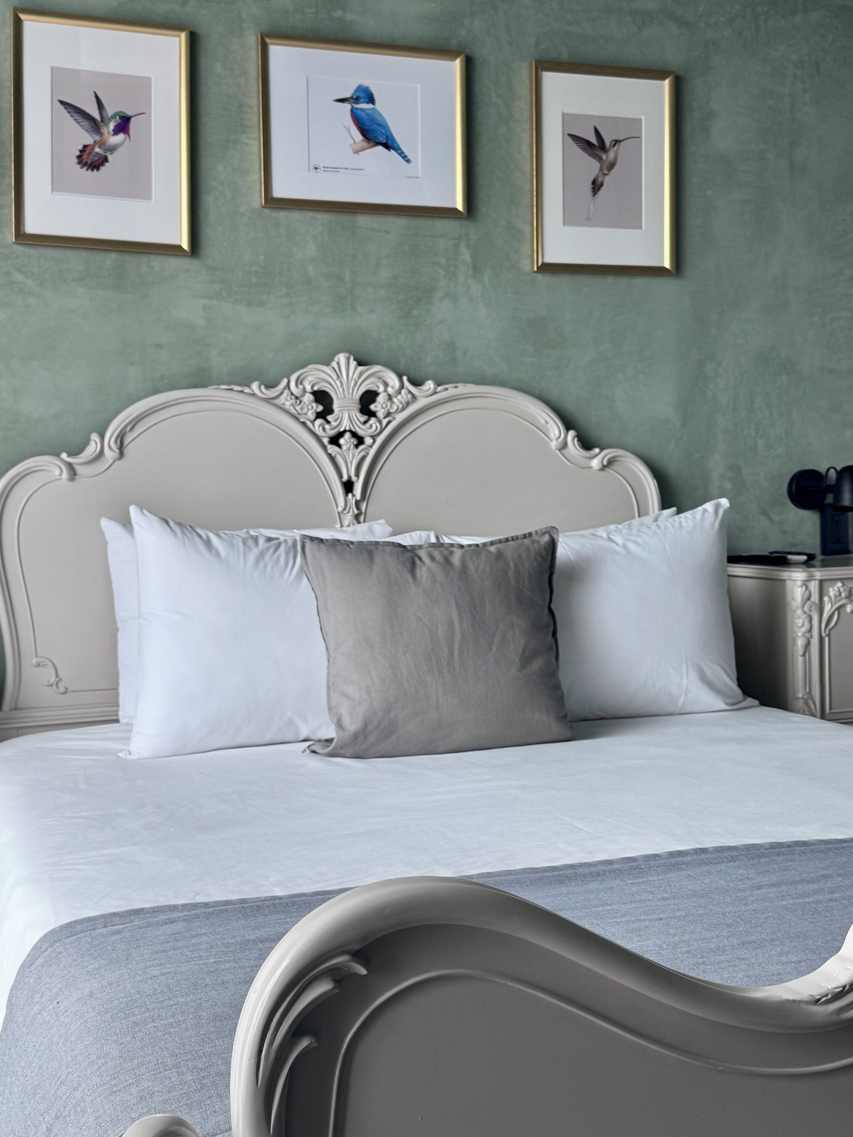 A bed with white and grey linens, a white ornate headboard, and three framed bird pictures on the wall.