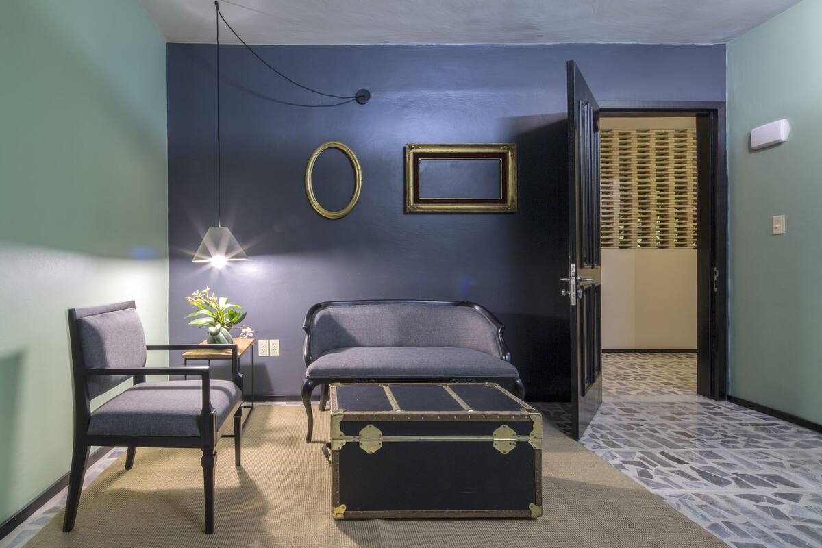 Een compacte kamer met een bed, een stoel, een klein tafeltje en een decoratieve koffer.