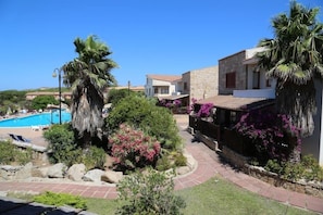 A pool area with a clear blue sky, palm trees, and a stone building with a red-tiled roof. - Club Esse Residence Capo D'orso (Palau)