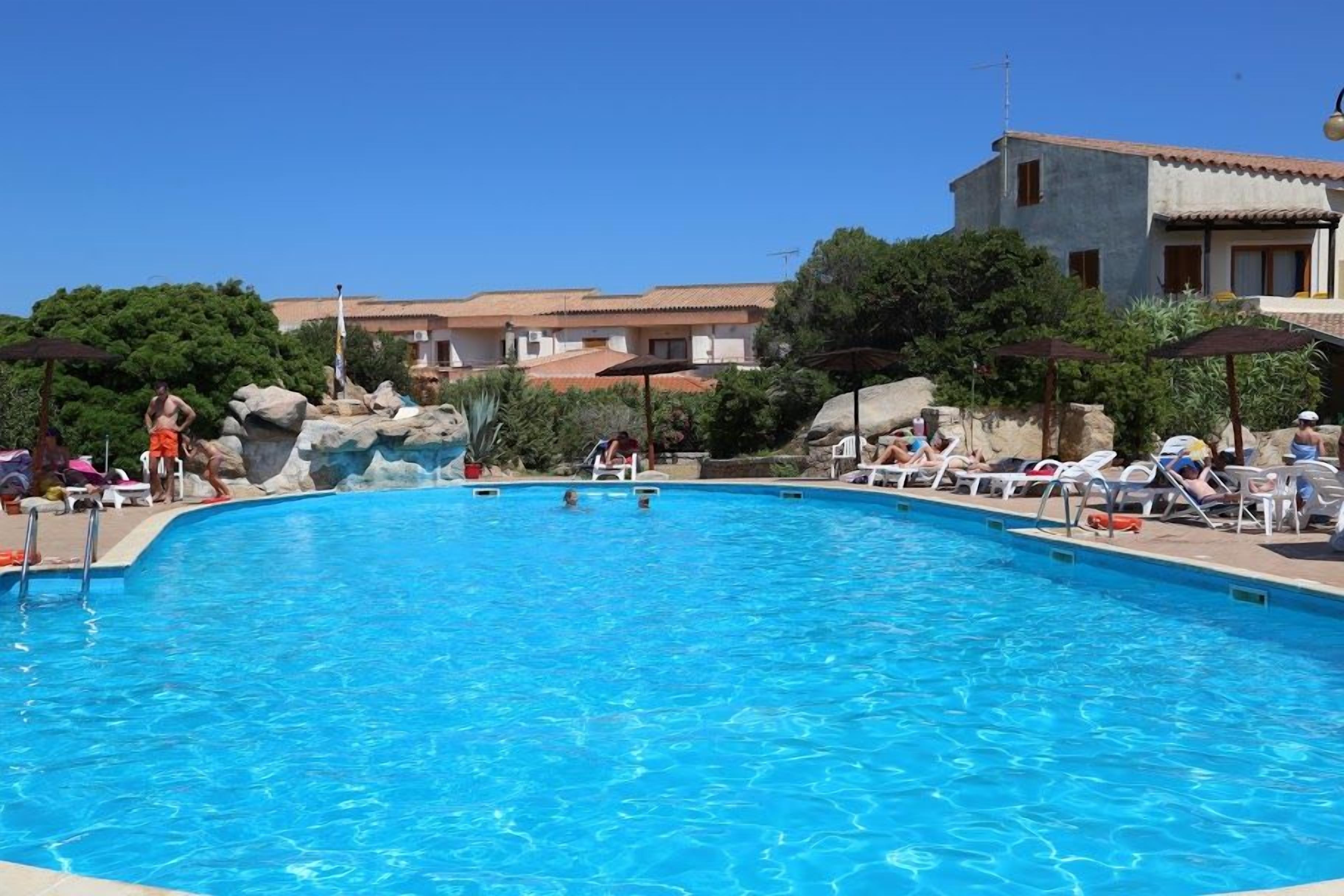 A swimming pool with lounge chairs and umbrellas, surrounded by greenery and buildings.