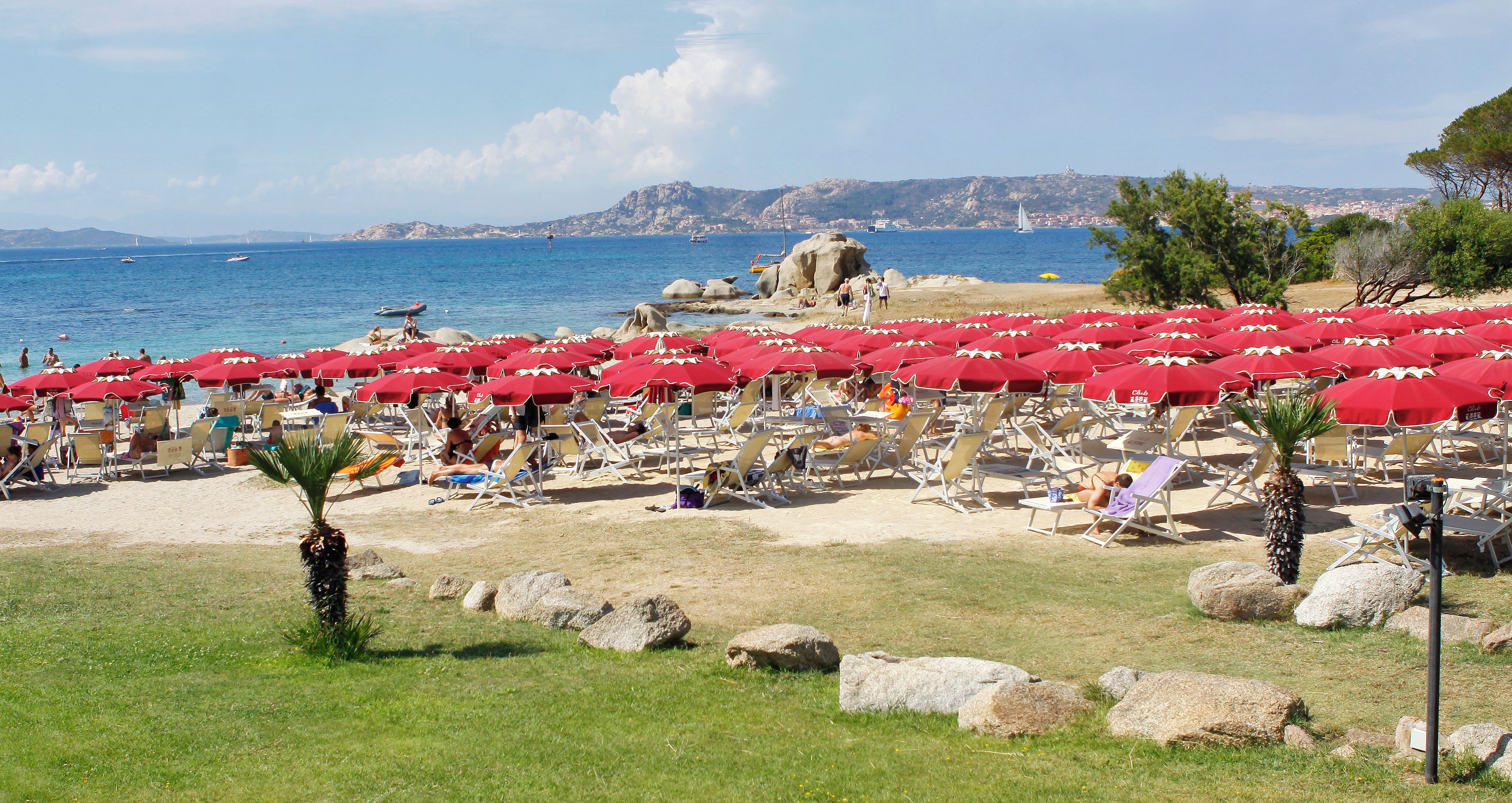 Una playa con hileras de sombrillas rojas y sillas de descanso, un mar azul cristalino y terreno rocoso al fondo.
