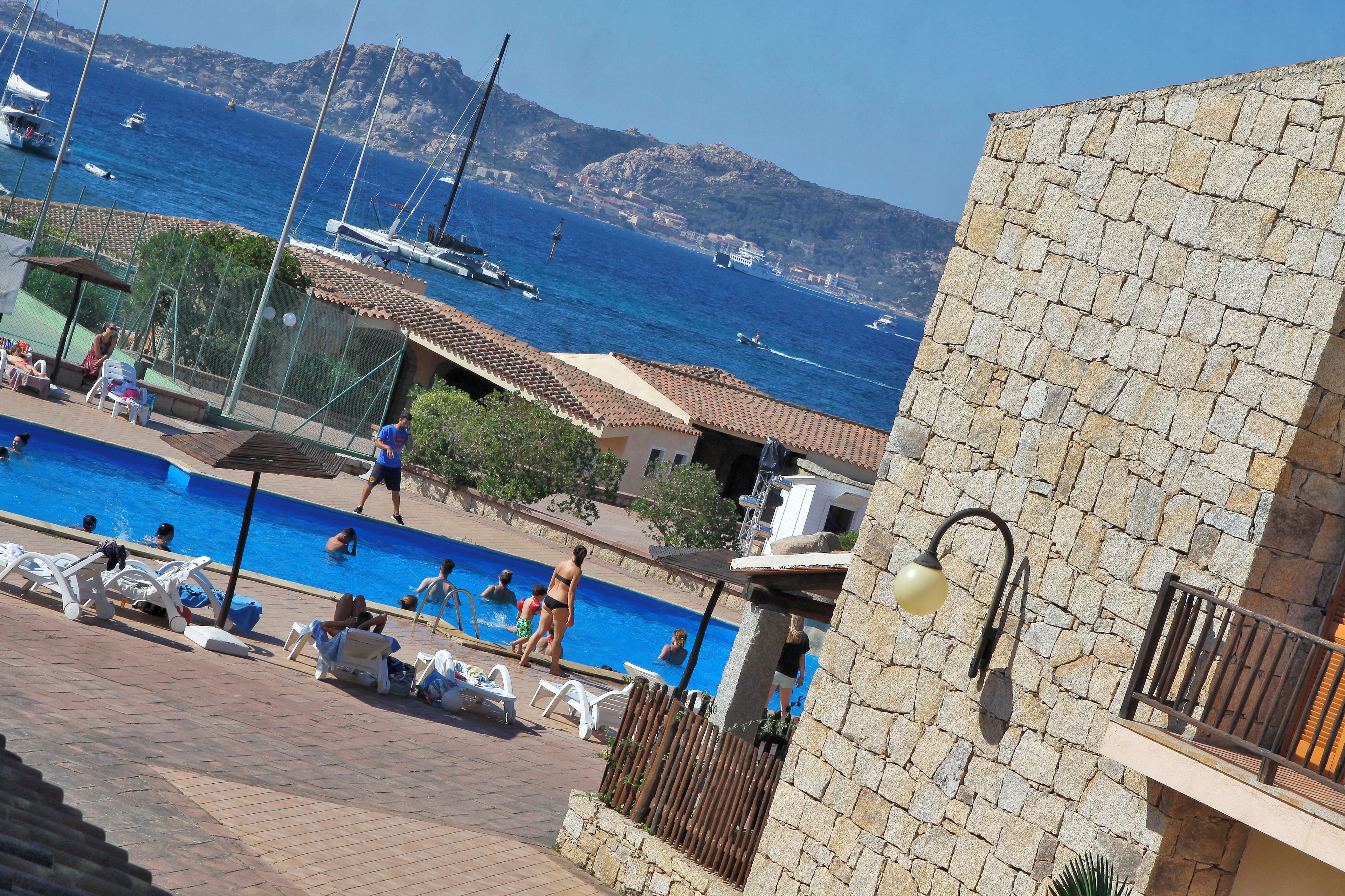 A swimming pool area with lounge chairs, a stone building, and boats in the background.