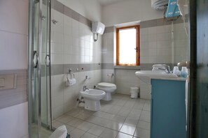 Ein Badezimmer mit Duschkabine aus Glas, Toilette, Bidet und Waschbecken mit Fenster darüber.