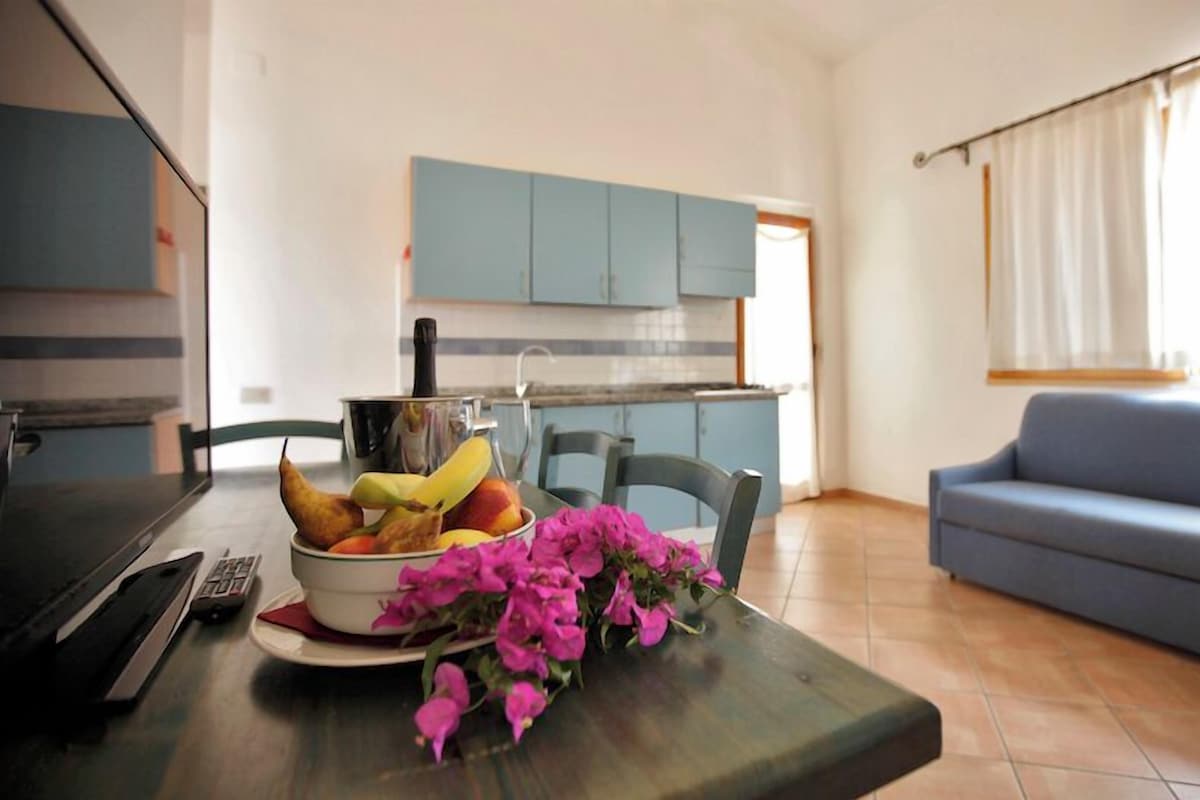 Un salon avec un canapé bleu, une table à manger ornée d’un saladier rempli de fruits et de fleurs, et une cuisine en arrière-plan.