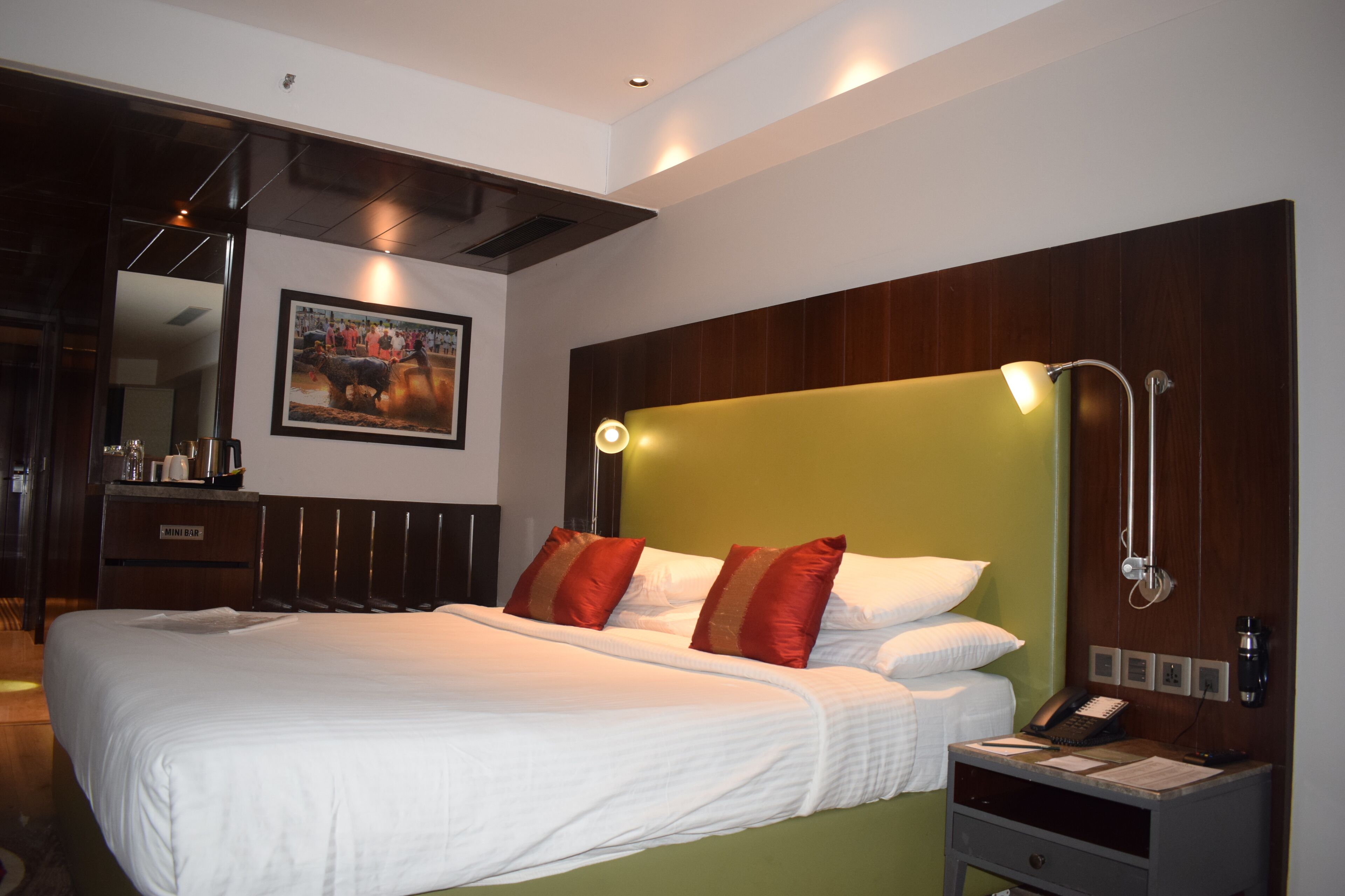 A hotel room with a large bed, a bedside table with a phone and papers, and a wall-mounted lamp.