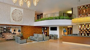 Un hall d’entrée spacieux avec un plafond haut, un grand lustre, un coin salon avec des canapés, un comptoir de réception et un balcon agrémenté de plantes.