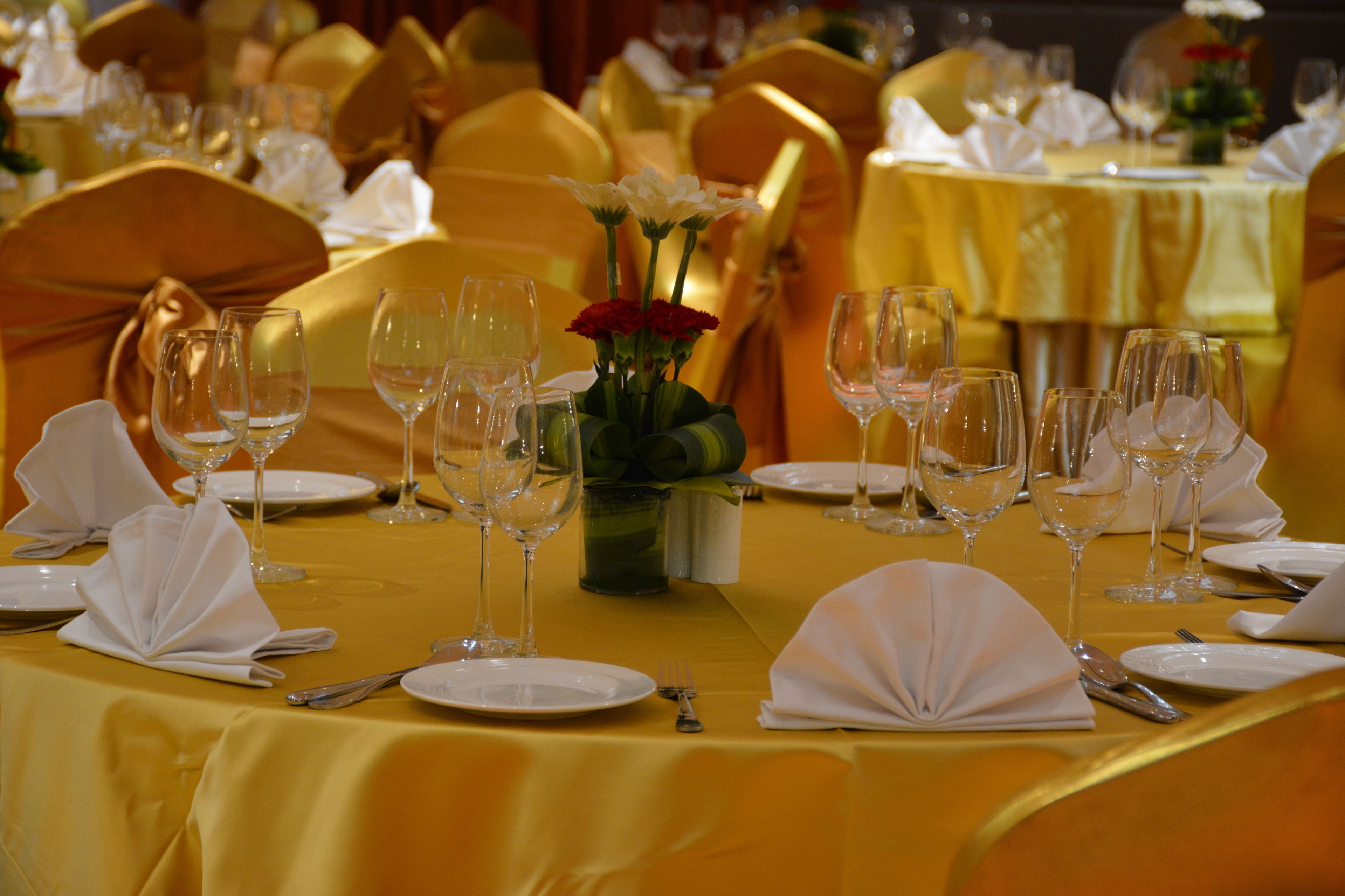 A banquet hall with tables set for a formal event, featuring white tablecloths, gold chairs, and empty wine glasses.