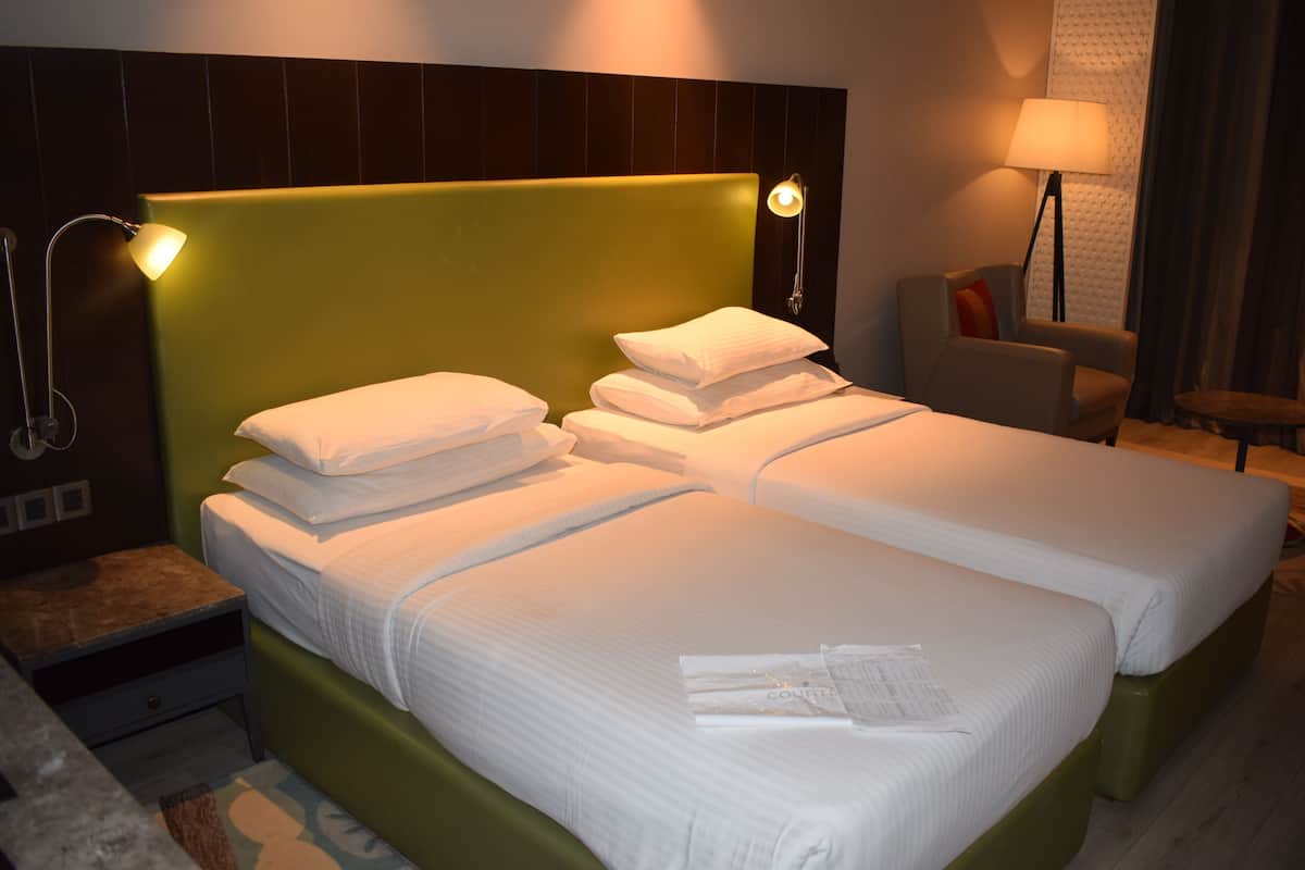 A hotel room with two beds, a green headboard, a nightstand, a lamp, and a chair.