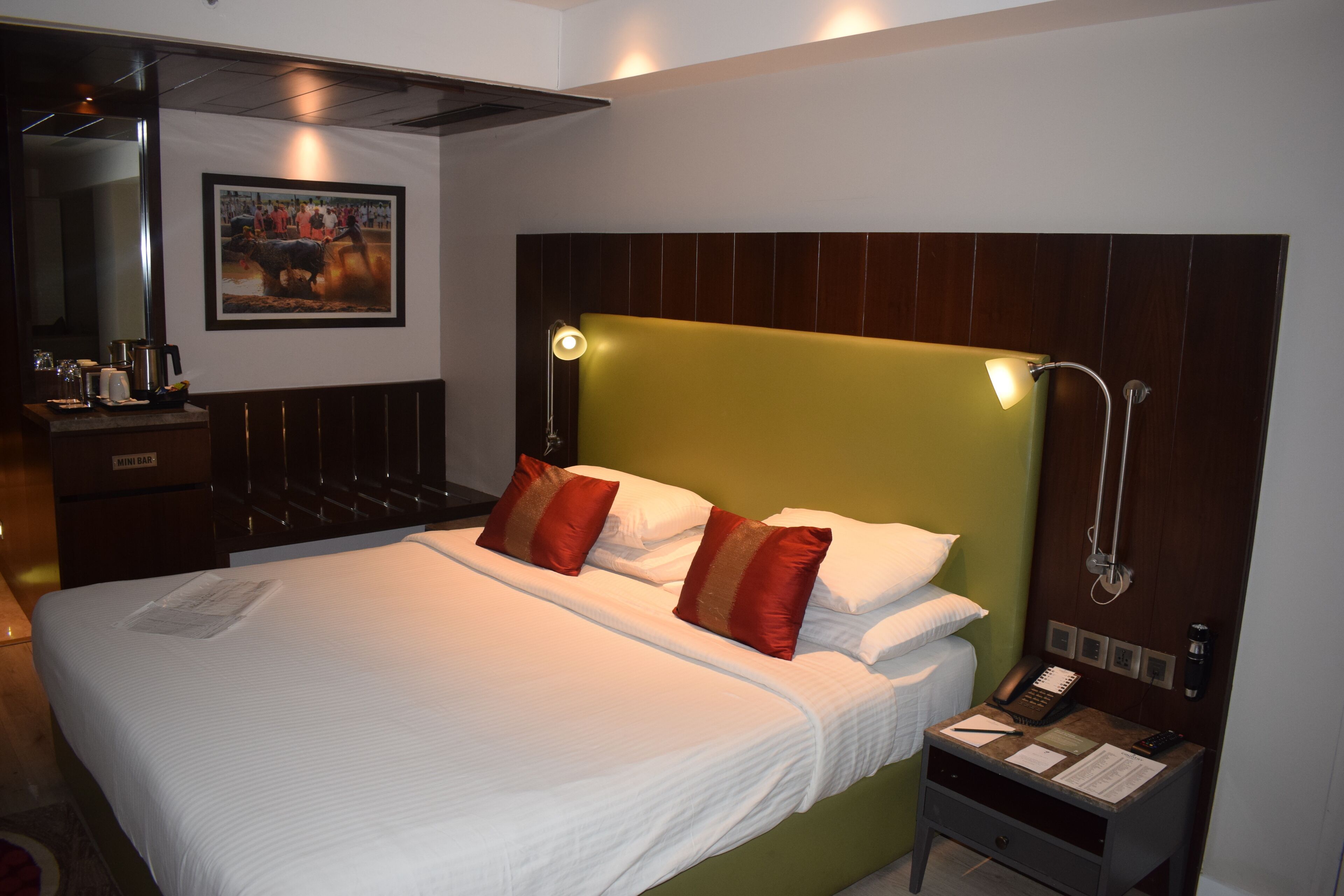 Habitación de hotel con una cama grande, una mesita de noche con un teléfono y documentos, un espejo y un cuadro en la pared.