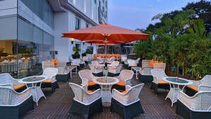 Une terrasse extérieure avec des meubles en osier et des coussins orange, un grand parasol rouge, et un bâtiment avec une enseigne indiquant « Country » en arrière-plan.