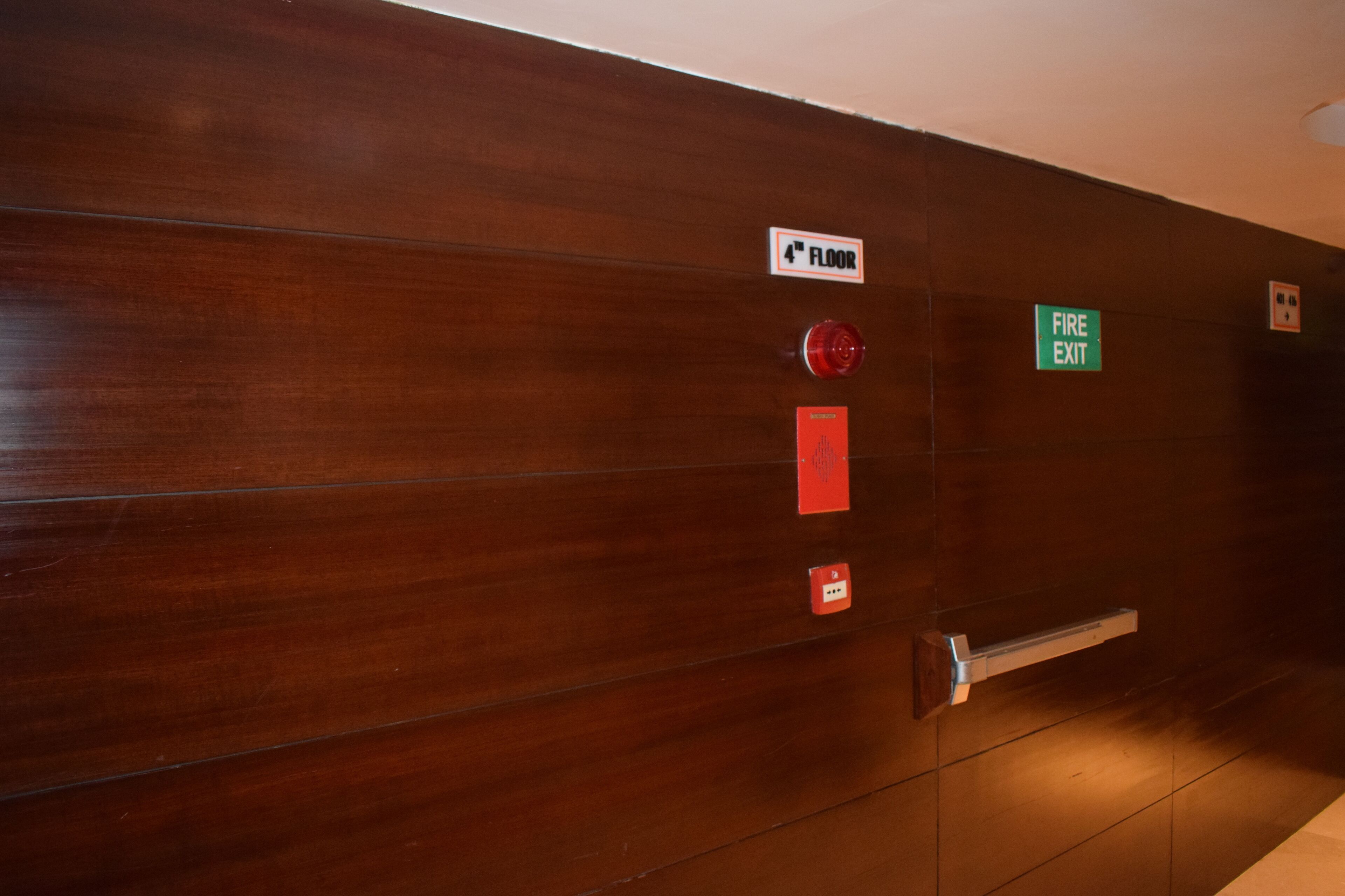 A wooden wall with a fire exit sign, a fire alarm, and an elevator control panel.