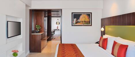 Una habitació d'hotel moderna amb un llit gran, un televisor de pantalla plana al mur, un quadre sobre el llit i una petita planta a la tauleta de nit.