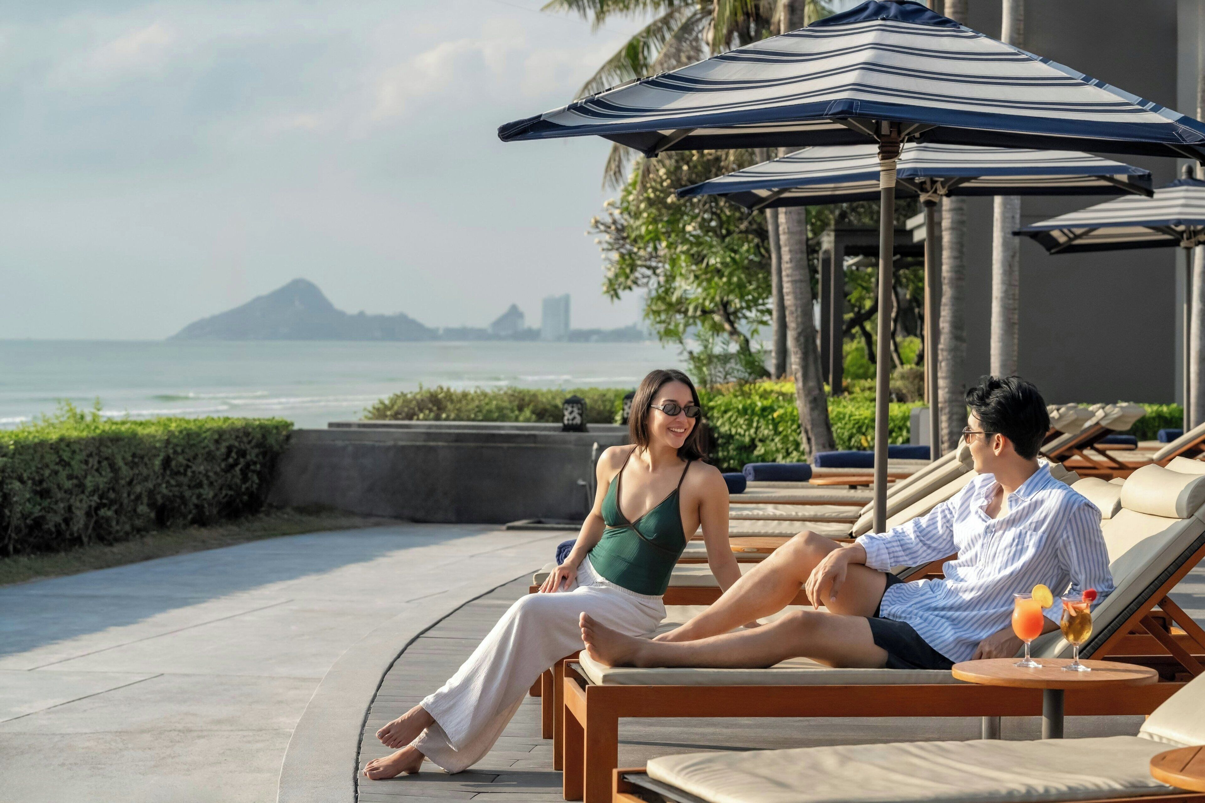 A couple relaxing on sun loungers by a pool with umbrellas, overlooking a body of water and distant land.