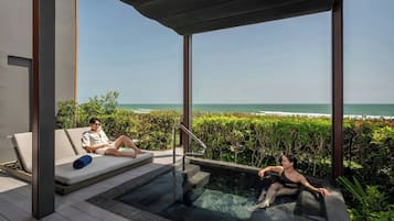 Un área de piscina moderna al aire libre con un jacuzzi de vidrio, una silla de descanso y vista al océano.