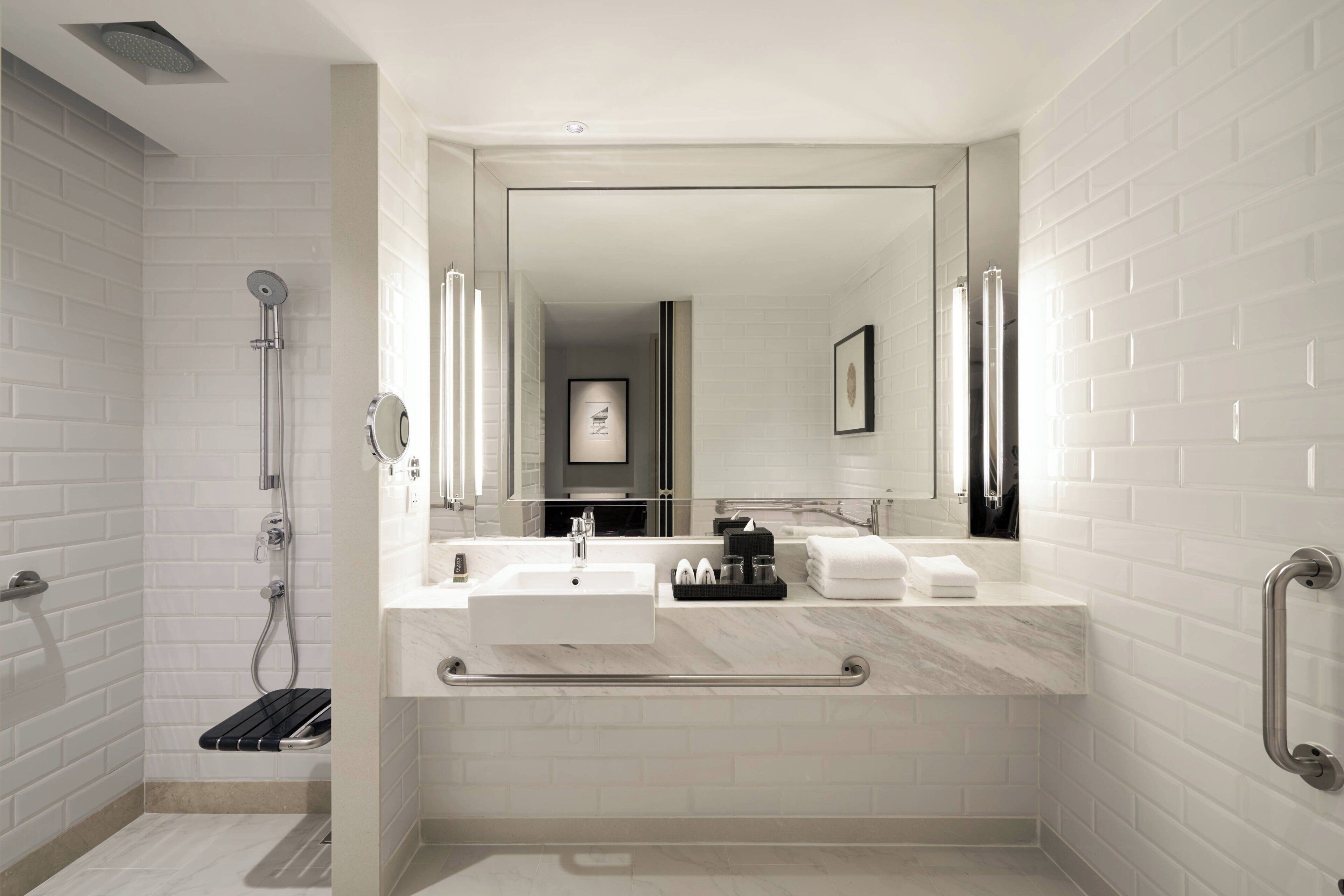 A modern bathroom with a walk-in shower, a large mirror, a sink, and a handrail.