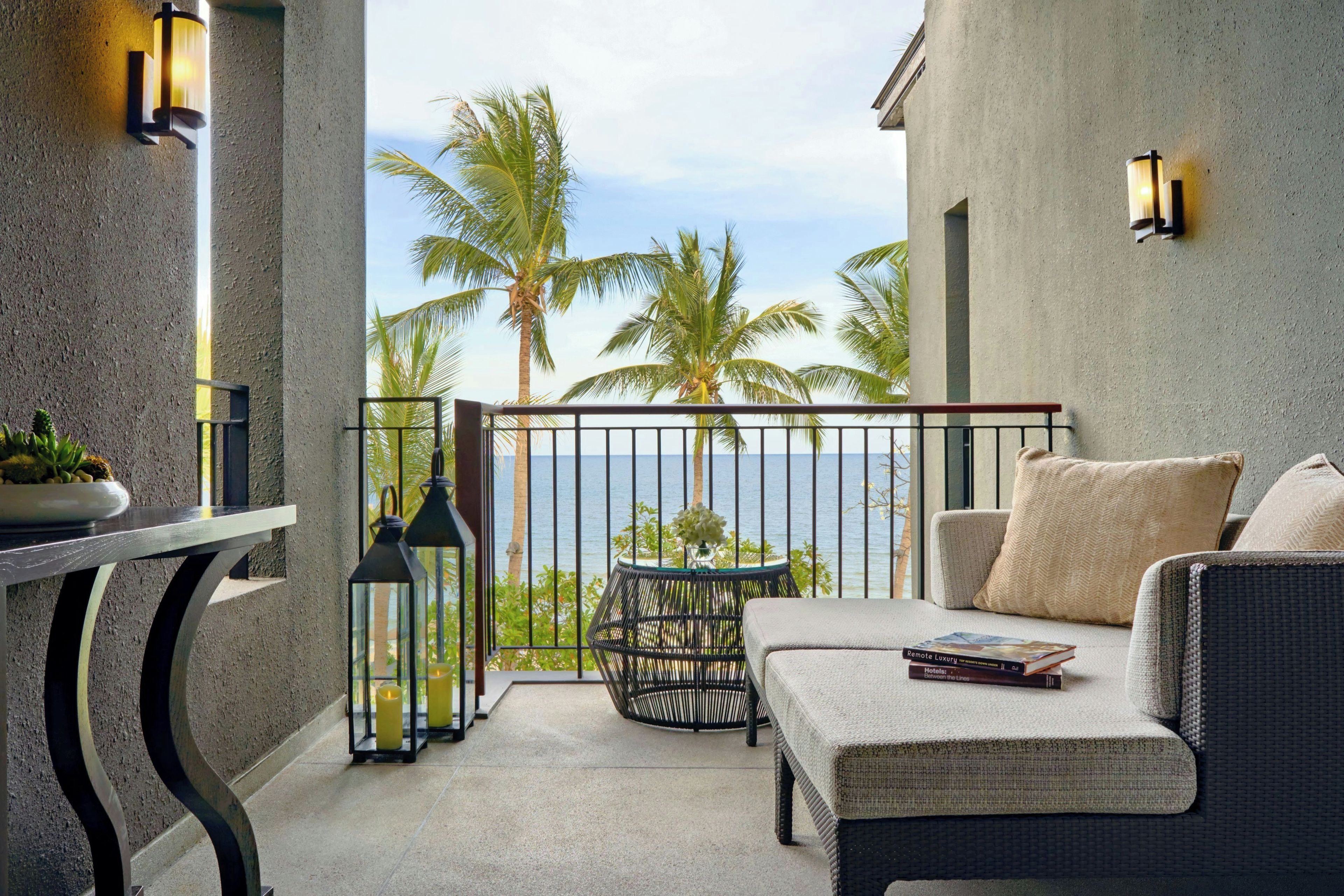 A balcony with a view of the sea, featuring a wicker sofa, a small table with a plant, and two wall-mounted lanterns.